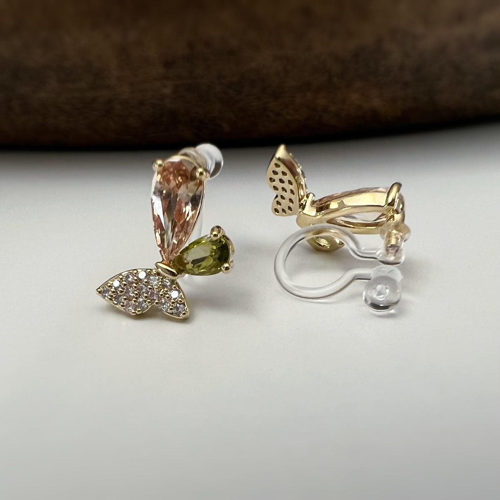 Clips d’oreilles papillon raffinés sertis de cristaux étincelants aux teintes champagne et vert olive. Un bijou délicat et lumineux.