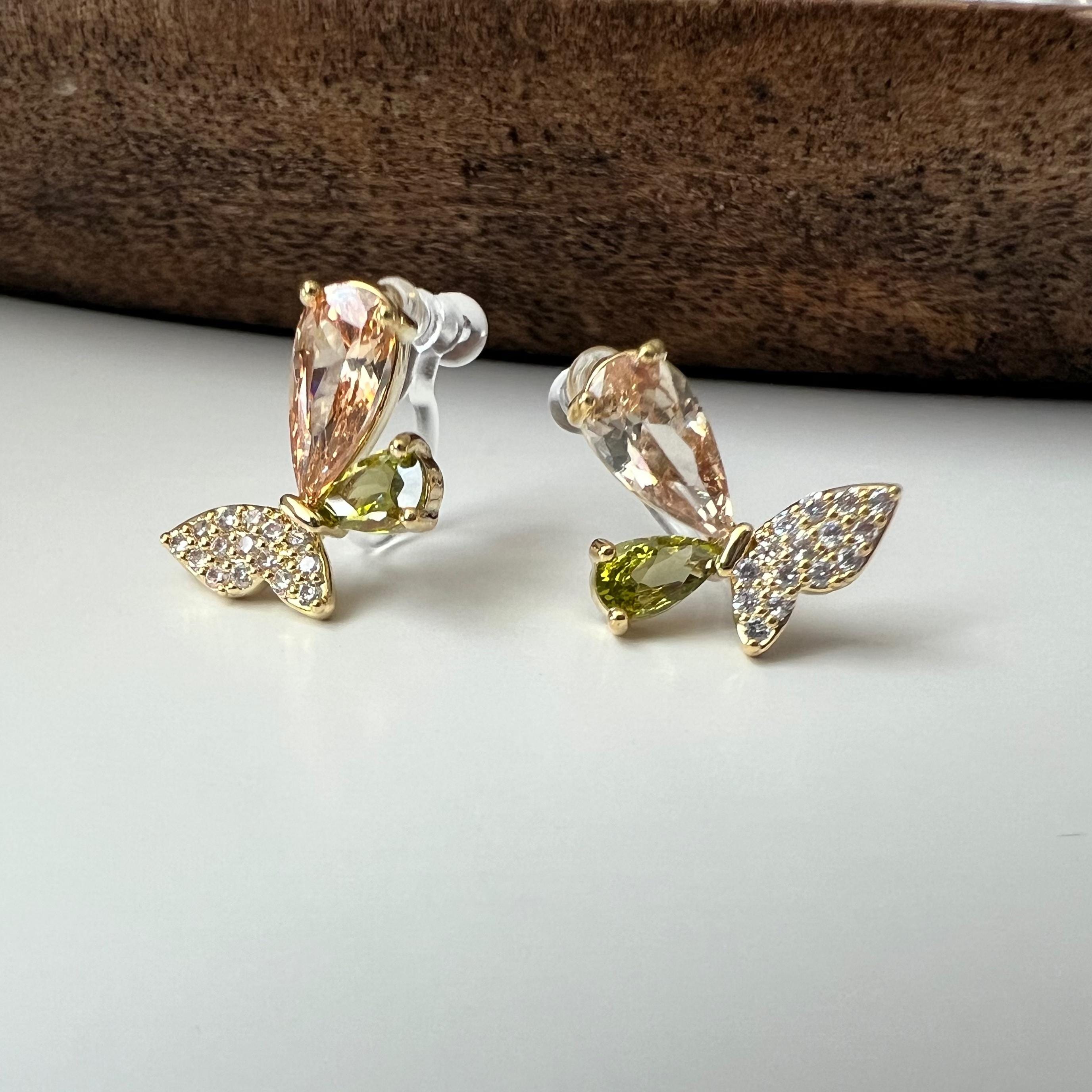 Clips d’oreilles papillon raffinés sertis de cristaux étincelants aux teintes champagne et vert olive. Un bijou délicat et lumineux.