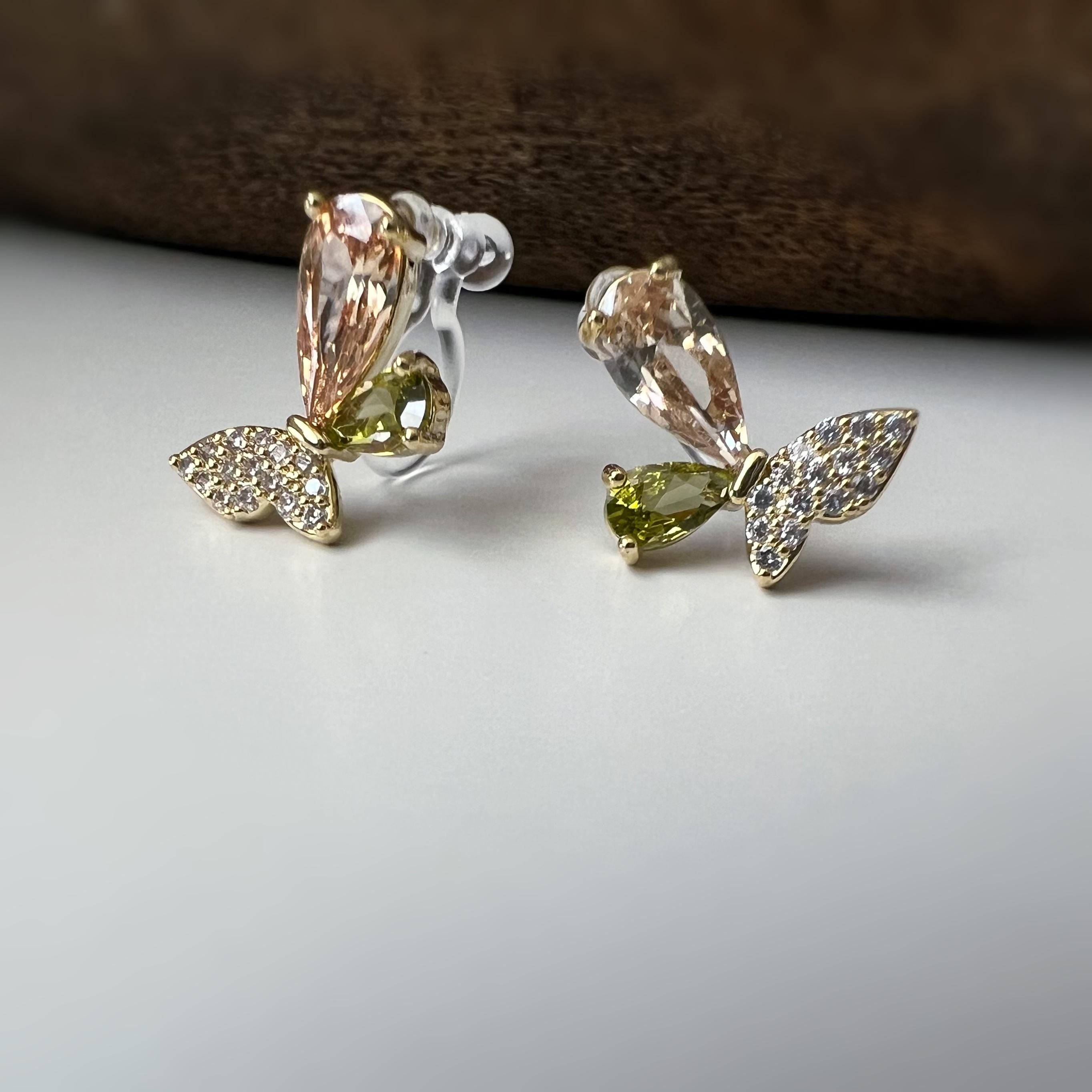 Clips d’oreilles papillon raffinés sertis de cristaux étincelants aux teintes champagne et vert olive. Un bijou délicat et lumineux.