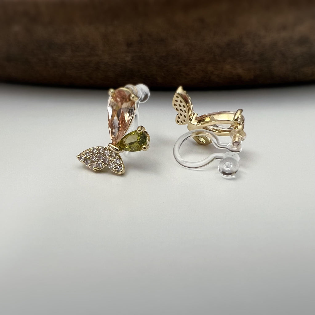 Clips d’oreilles papillon raffinés sertis de cristaux étincelants aux teintes champagne et vert olive. Un bijou délicat et lumineux.