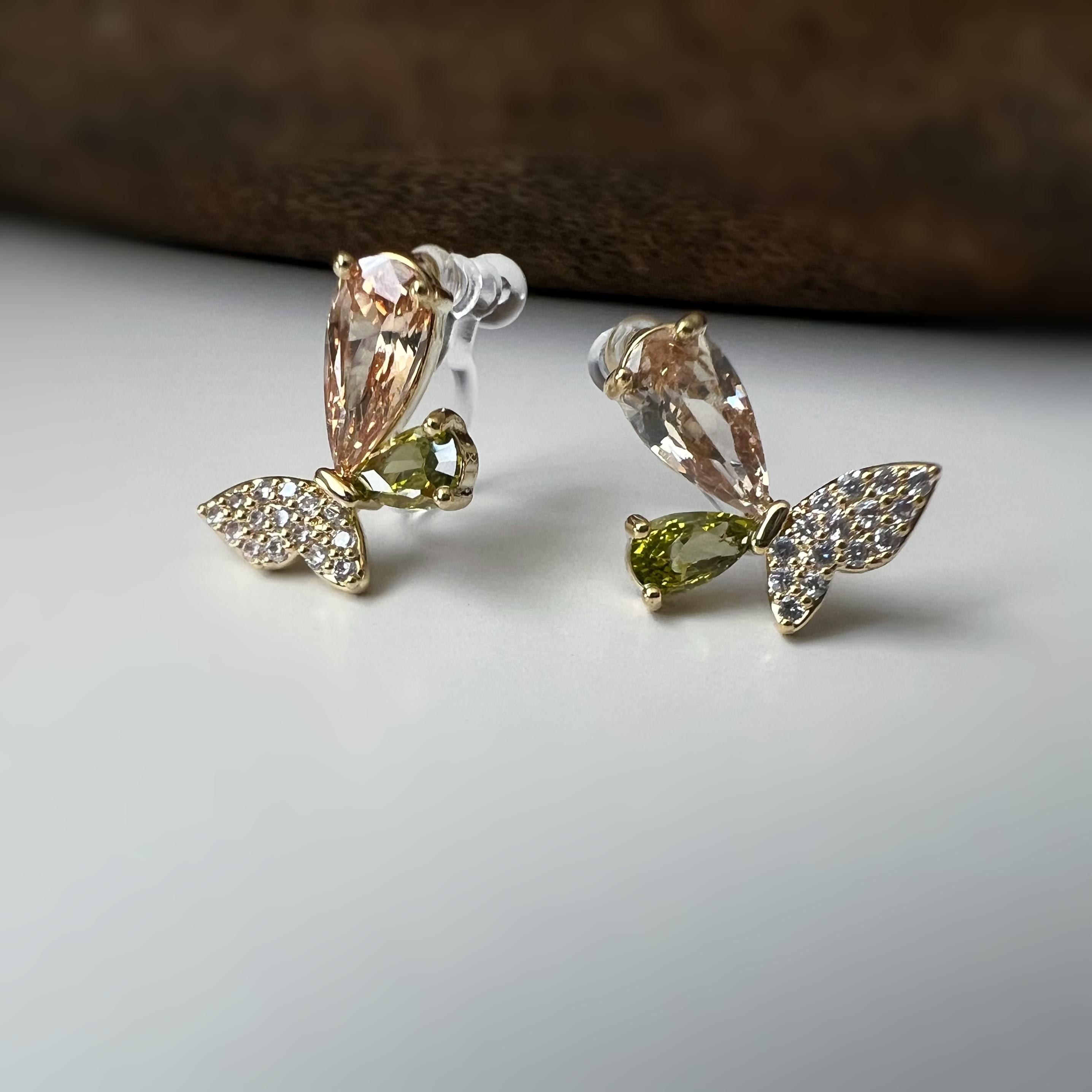 Clips d’oreilles papillon raffinés sertis de cristaux étincelants aux teintes champagne et vert olive. Un bijou délicat et lumineux.